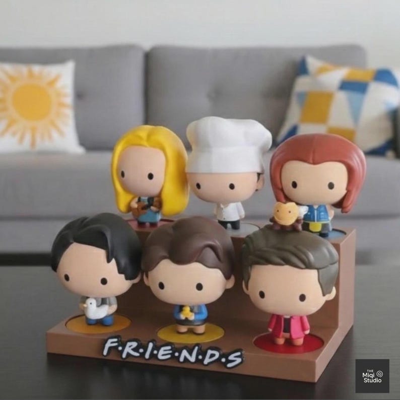 May include: A collection of six cartoon character figurines from the TV show "Friends" displayed on a brown stand. The figurines have large heads and small bodies, with the show's logo "F.R.I.E.N.D.S" printed on the base.