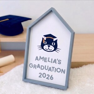 May include: A framed sign with a gray house-shaped frame. The sign reads "Amelia's Graduation 2026" below a cat wearing a graduation cap. A navy blue graduation cap with a gold tassel sits on a shelf in the background.