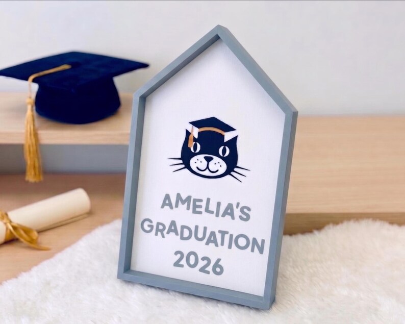 May include: A framed sign with a gray house-shaped frame. The sign reads "Amelia's Graduation 2026" below a cat wearing a graduation cap. A navy blue graduation cap with a gold tassel sits on a shelf in the background.