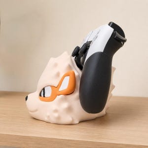 May include: A cream-colored, cartoon-style controller holder shaped like a fox. The fox has orange and white accents around the eyes and ears. A black and white game controller is placed in the holder, with the black grip visible.