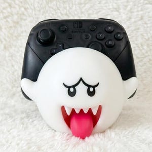May include: A black Nintendo Switch Pro controller mounted on a white, spherical Boo character stand. The stand features the ghost's signature wide eyes, sharp white teeth, and a protruding pink tongue, with the controller buttons visible above.