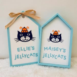 Puede incluir: Dos letreros en forma de casa de color azul claro con centros blancos. Cada letrero presenta una ilustraci&oacute;n de un gato negro con un sombrero naranja y el texto "ELLIE'S JELLYCATS" y "MAISEY'S JELLYCATS" en azul claro. El letrero izquierdo tiene un lazo beige.