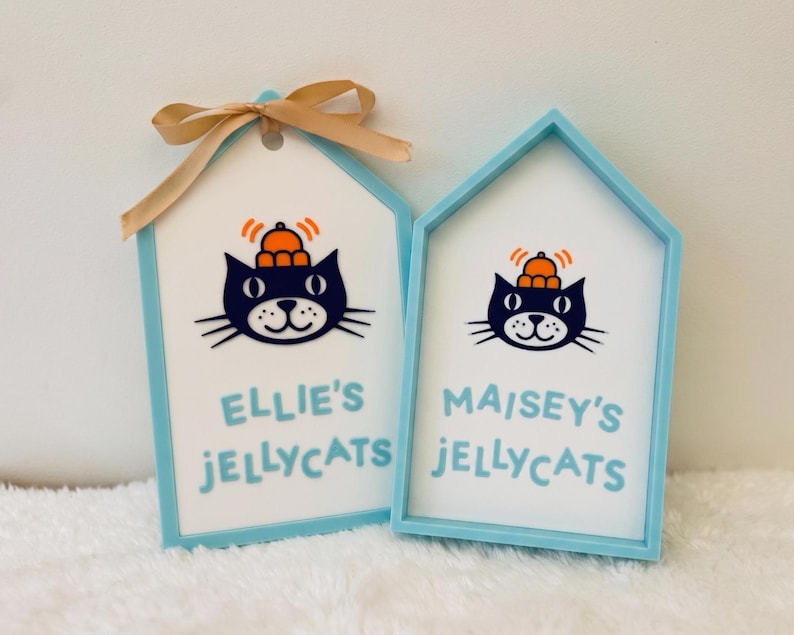 Puede incluir: Dos letreros en forma de casa de color azul claro con centros blancos. Cada letrero presenta una ilustraci&oacute;n de un gato negro con un sombrero naranja y el texto "ELLIE'S JELLYCATS" y "MAISEY'S JELLYCATS" en azul claro. El letrero izquierdo tiene un lazo beige.