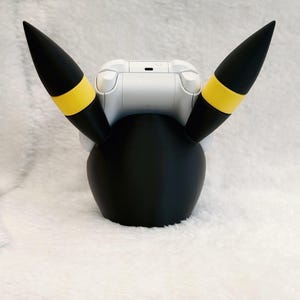May include: A black controller stand featuring two tall, pointed ears with yellow bands, designed to hold a white game controller. The stand has a smooth, rounded base and the ears extend upward, mimicking the appearance of a creature's head.