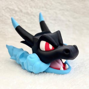 May include: A stylized black dragon head figurine with bright red eyes and a wide-open mouth showing white teeth and a pink tongue. The creature features two black horns with light blue tips and a light blue cloud-like base surrounding its neck.