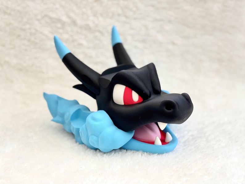 May include: A stylized black dragon head figurine with bright red eyes and a wide-open mouth showing white teeth and a pink tongue. The creature features two black horns with light blue tips and a light blue cloud-like base surrounding its neck.