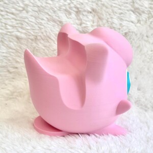 Peut inclure: Un support de t&eacute;l&eacute;phone rose clair imprim&eacute; en 3D en forme de personnage rond avec une finition mate lisse. Le support pr&eacute;sente une rainure profonde et incurv&eacute;e sur le dessus pour maintenir un appareil mobile et une petite base plate.
