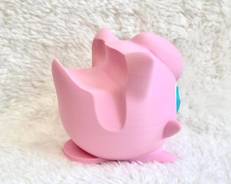 Peut inclure: Un support de t&eacute;l&eacute;phone rose clair imprim&eacute; en 3D en forme de personnage rond avec une finition mate lisse. Le support pr&eacute;sente une rainure profonde et incurv&eacute;e sur le dessus pour maintenir un appareil mobile et une petite base plate.
