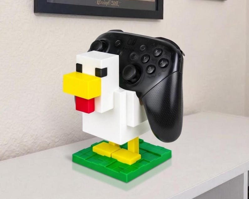 May include: A white, yellow, red, and black chicken-shaped game controller stand. The stand holds a black game controller. The chicken has a yellow beak, red tongue, and black eyes. The base is green with yellow feet.