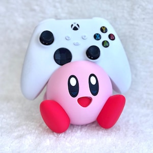 May include: A white Xbox wireless controller resting on a spherical pink Kirby figure with red feet and a smiling face. The controller features black analogue sticks, a black directional pad, and multicoloured action buttons including Y, B, A, and X.