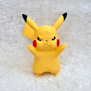 May include: A yellow Pikachu figurine with a grumpy facial expression, featuring pointed ears with black tips, round red cheek patches, and small arms raised in a defensive posture. The character has a smooth matte finish and a lightning bolt tail.
