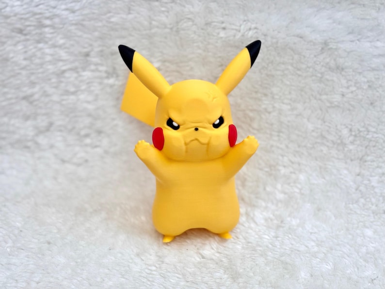 May include: A yellow Pikachu figurine with a grumpy facial expression, featuring pointed ears with black tips, round red cheek patches, and small arms raised in a defensive posture. The character has a smooth matte finish and a lightning bolt tail.