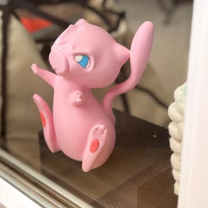 May include: A pink, stylised creature figurine with a long, slender tail, pointed ears, and large, expressive blue eyes. The figure is depicted in a dynamic, upright pose with small arms extended and feet positioned forward, featuring small red accents.