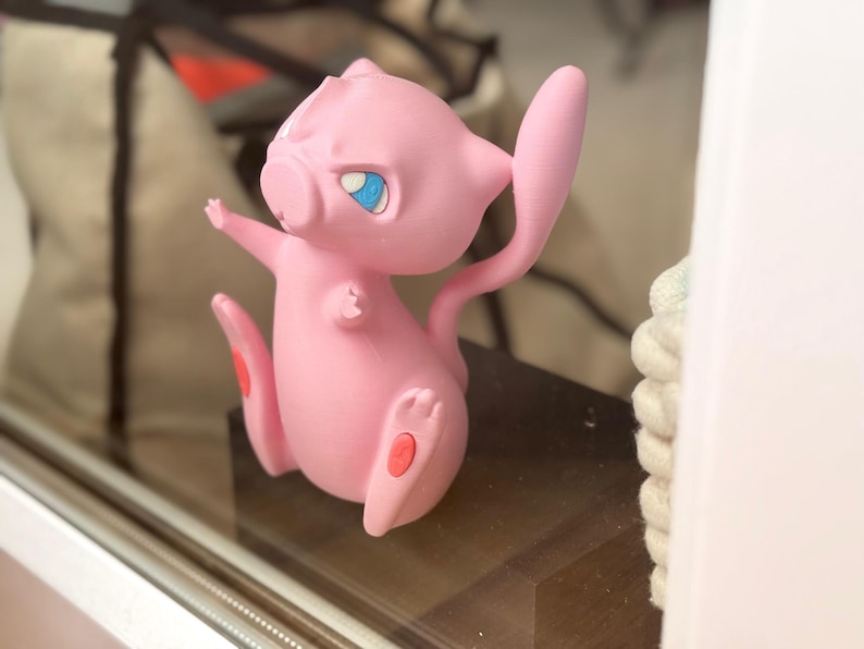May include: A pink, stylised creature figurine with a long, slender tail, pointed ears, and large, expressive blue eyes. The figure is depicted in a dynamic, upright pose with small arms extended and feet positioned forward, featuring small red accents.