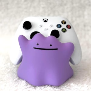 Puede incluir: Una figura de personaje con forma de mancha morada con una línea negra simple para la boca y dos puntos negros para los ojos, que sirve como soporte para un controlador de videojuegos inalámbrico blanco con joysticks negros y botones de colores.