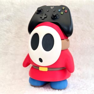 May include: A red and blue character figure with a white oval mask featuring two black eye holes and a small black mouth. The figure wears a brown belt with a yellow buckle and supports a black wireless game controller on its head.
