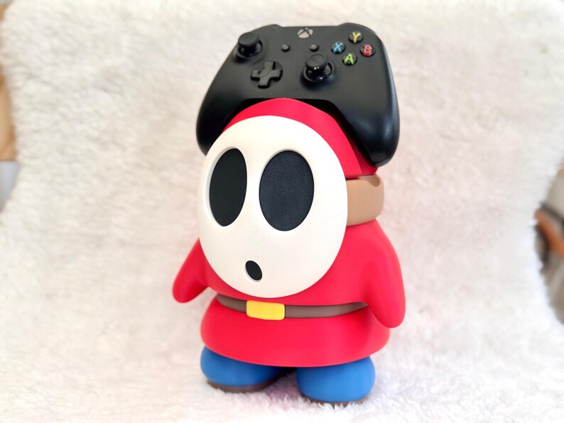 May include: A red and blue character figure with a white oval mask featuring two black eye holes and a small black mouth. The figure wears a brown belt with a yellow buckle and supports a black wireless game controller on its head.