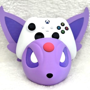 May include: A white video game controller rests inside a purple and lavender creature-shaped stand. The stand features large pointed ears, a rounded head with a small red circular accent on the forehead, and two white eye-shaped cutouts.