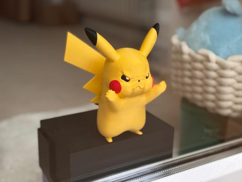 May include: A yellow Pikachu figurine with a determined facial expression, featuring pointed ears with black tips, red circular cheeks, and a jagged lightning bolt-shaped tail. The character is standing in a dynamic pose with arms raised.
