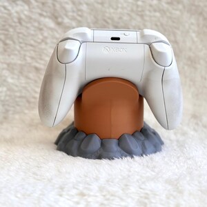 May include: A white Xbox wireless controller resting on a brown cylindrical stand with a gray, rock-textured base. The controller features the Xbox logo on the top center, with two shoulder buttons and a charging port visible on the back panel.