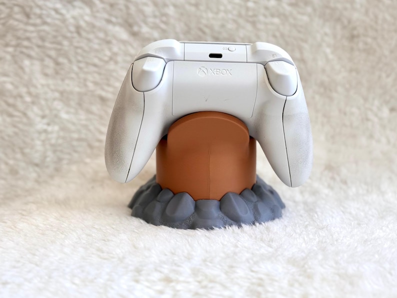May include: A white Xbox wireless controller resting on a brown cylindrical stand with a gray, rock-textured base. The controller features the Xbox logo on the top center, with two shoulder buttons and a charging port visible on the back panel.