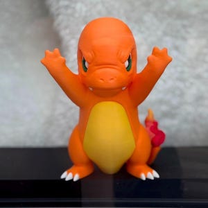 May include: An orange and yellow bipedal creature figurine with a rounded head, green eyes, and a small mouth. The figure has raised arms with three-fingered hands, white claws on its feet, and a red flame-shaped tail extending from its back.