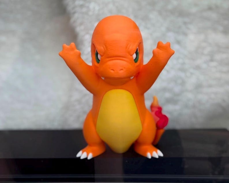 May include: An orange and yellow bipedal creature figurine with a rounded head, green eyes, and a small mouth. The figure has raised arms with three-fingered hands, white claws on its feet, and a red flame-shaped tail extending from its back.