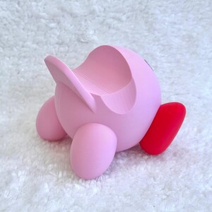 May include: A pink, spherical character-shaped stand featuring a smooth, matte finish with a curved indentation on the top for holding items. The object has two rounded pink feet and one prominent, bright red foot extending to the side.