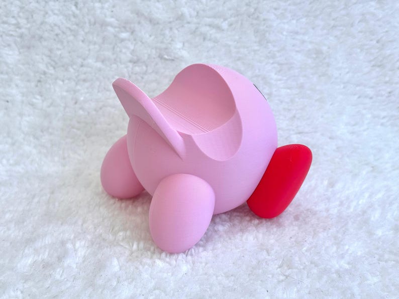 May include: A pink, spherical character-shaped stand featuring a smooth, matte finish with a curved indentation on the top for holding items. The object has two rounded pink feet and one prominent, bright red foot extending to the side.