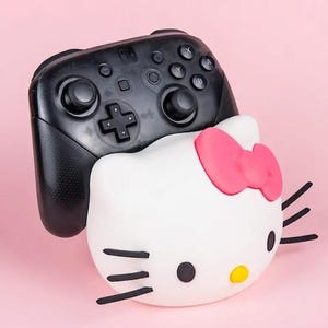 May include: A black Nintendo Switch Pro Controller rests on top of a white Hello Kitty-shaped accessory with a pink bow. The accessory has black whiskers and a yellow nose. The background is a solid pink.