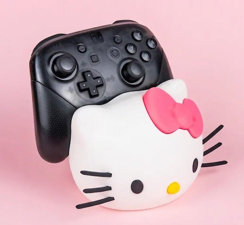May include: A black Nintendo Switch Pro Controller rests on top of a white Hello Kitty-shaped accessory with a pink bow. The accessory has black whiskers and a yellow nose. The background is a solid pink.