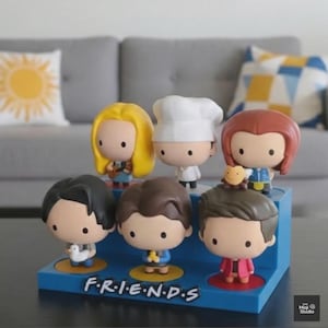 May include: A set of seven cartoon figurines of characters from the TV show "Friends". The figurines have large heads and small bodies, with distinctive hairstyles and outfits. They are displayed on a blue base with the show's logo.