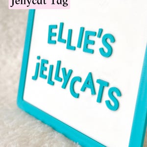 Puede incluir: Un letrero rectangular con un borde turquesa brillante y un centro blanco. El letrero presenta letras turquesas en relieve que dicen "ELLIE'S JELLYCATS" y un gr&aacute;fico negro de una cara de gato en la parte superior. Se ve una etiqueta rosa "Jellycat Tag".