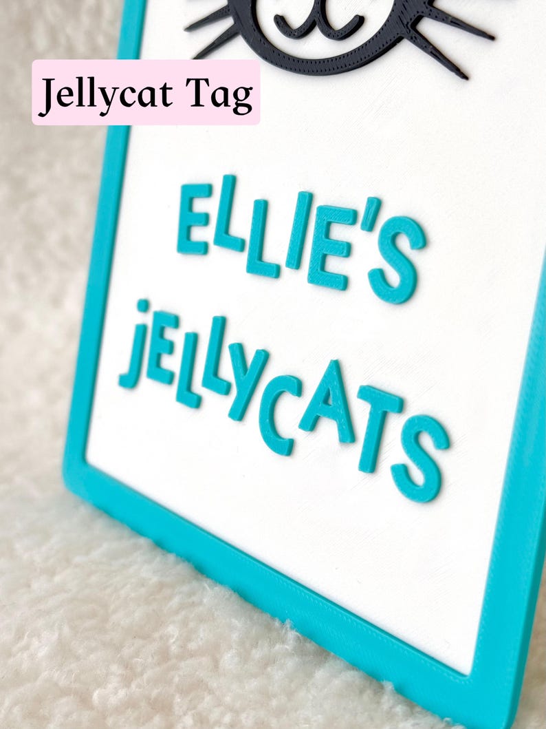 Puede incluir: Un letrero rectangular con un borde turquesa brillante y un centro blanco. El letrero presenta letras turquesas en relieve que dicen "ELLIE'S JELLYCATS" y un gr&aacute;fico negro de una cara de gato en la parte superior. Se ve una etiqueta rosa "Jellycat Tag".