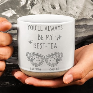 May include: White ceramic mug with the text "YOU'LL ALWAYS BE MY BEST-TEA" and two smiling teacup illustrations. The names "SIENNA" and "CALLIE" are printed below the teacups. The mug has a grey handle and a tan base.