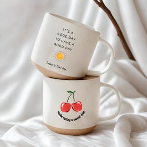 May include: Two white ceramic mugs with a speckled finish. One mug has the text "IT'S A GOOD DAY TO HAVE A GOOD DAY" with a sun graphic. The other mug has a cherry graphic and the text "make today a sweet day".