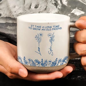 Pode incluir: Uma caneca de cerâmica branca com base e alça bege. A caneca apresenta ilustrações florais azuis e o texto "IT TAKE A LONG TIME TO GROW AN OLD FRIEND" com os nomes "Luna" e "Sophie".