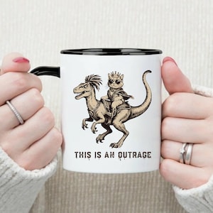 以下が含まれることがあります： 黒い縁とハンドルが付いた白いセラミックマグ。マグには、王冠をかぶった猫が恐竜に乗っているセピア調のイラストと、その下に「THIS IS AN OUTRAGE」というテキストが描かれています。