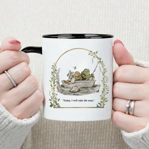 May include: White ceramic mug with a black rim and handle. The mug features an illustration of two frogs in a boat, with the text "Today, I will take life easy." The mug is held by a person.