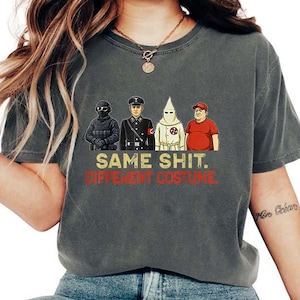 May include: A dark grey t-shirt featuring the text "SAME SHIT. DIFFERENT COSTUME." and illustrations of four figures in different uniforms. The t-shirt is a casual style, suitable for everyday wear.