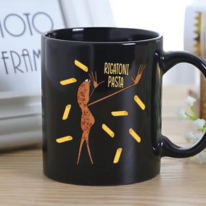 Puede incluir: Taza de cerámica negra con una ilustración humorística. Presenta una figura de pasta de dibujos animados con los brazos levantados, rodeada de formas de pasta amarillas y el texto "RIGATONI PASTA". La taza tiene un asa curvada.