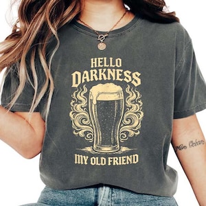 May include: Dark gray t-shirt with the text "HELLO DARKNESS MY OLD FRIEND" in a vintage style. The graphic features a beer glass with foam and decorative swirls. The shirt is a casual wear item.