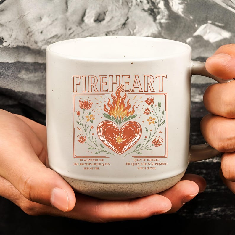 K&ouml;nnte beinhalten: Cremefarbene Keramik-Tasse mit dem Wort "FIREHEART" in orangefarbenen Buchstaben. Die Tasse zeigt ein Herzdesign mit Flammen und floralen Akzenten. Der Boden der Tasse ist aus dunklerer, unglasierter Keramik.