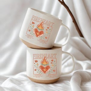 K&ouml;nnte beinhalten: Zwei wei&szlig;e Keramikbecher &uuml;bereinander gestapelt. Jeder Becher hat das Wort "FIREHEART" &uuml;ber einem Herzen mit Flammen, umgeben von floralen Illustrationen. Die Becher haben einen beigefarbenen Boden und einen wei&szlig;en Henkel.