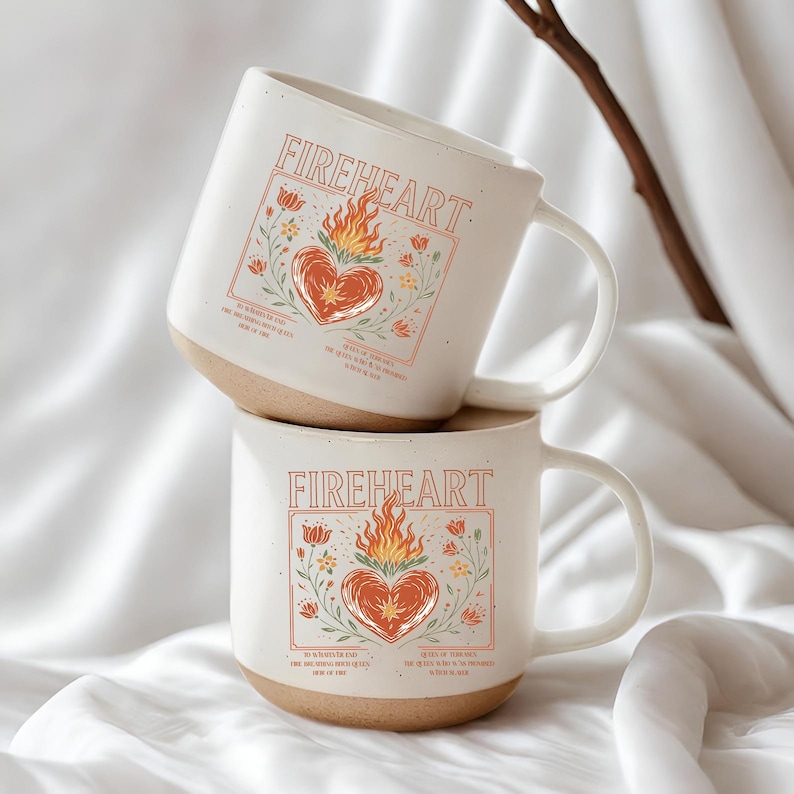 K&ouml;nnte beinhalten: Zwei wei&szlig;e Keramikbecher &uuml;bereinander gestapelt. Jeder Becher hat das Wort "FIREHEART" &uuml;ber einem Herzen mit Flammen, umgeben von floralen Illustrationen. Die Becher haben einen beigefarbenen Boden und einen wei&szlig;en Henkel.