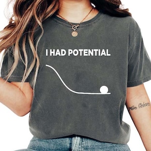 Könnte beinhalten: Dunkelgraues T-Shirt mit dem weißen Text "I HAD POTENTIAL". Unter dem Text befindet sich eine weiße Grafik einer gebogenen Linie und einer kleinen weißen Kugel. Das Shirt ist ein lässiger Unisex-Stil.