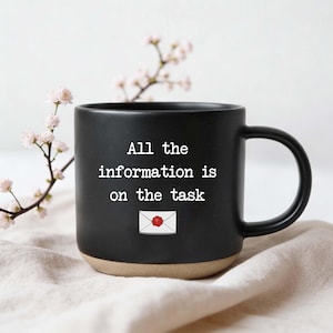 May include: A black ceramic coffee mug with a beige base. The mug features the text "All the information is on the task" in white, along with a small envelope graphic. A sprig of pink flowers is in the background.