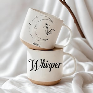 Puede incluir: Dos tazas de cerámica blancas apiladas con base beige. La taza superior tiene un diseño de luna creciente negra y floral con la palabra "Aries". La taza inferior tiene la palabra "Whisper" en escritura negra.