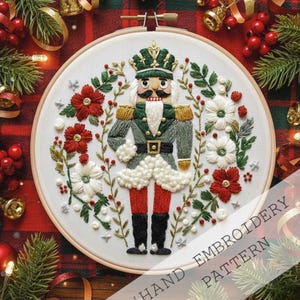 May include: Hand-embroidered Christmas-themed hoop art featuring a nutcracker soldier. The design includes a green and gold uniform, red pants, and black boots, surrounded by red and white flowers and greenery. The text "HAND EMBROIDERY PATTERN" is visible.