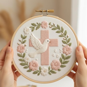 May include: Embroidered hoop art featuring a white dove, a pink cross, and floral accents. The design is set against a white fabric background, framed by a wooden hoop. The embroidery includes pink and white flowers and green leaves.